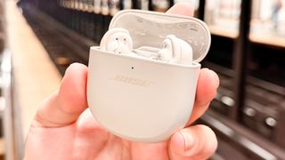 Listing image for best noise-cancelling earbuds round up showing Bose QuietComfort Ultra Earbuds in charging case held in hand by with a blurred out subway track in background